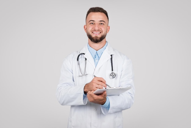 Doctor hombre sonriente con estetoscopio escribiendo en el portapapeles