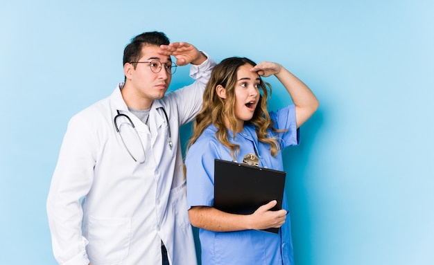 Doctor joven pareja posando en una pared azul aislado mirando lejos manteniendo la mano en la frente.