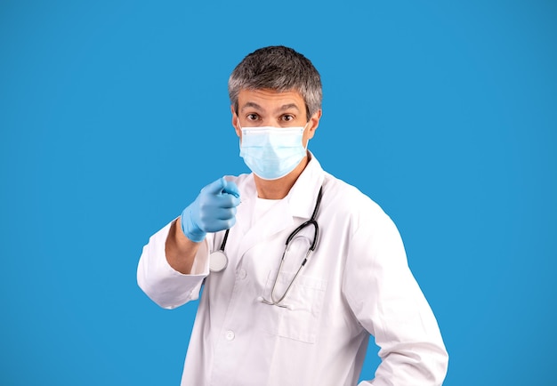 Doctor en máscara facial apuntando a la cámara sobre fondo azul.