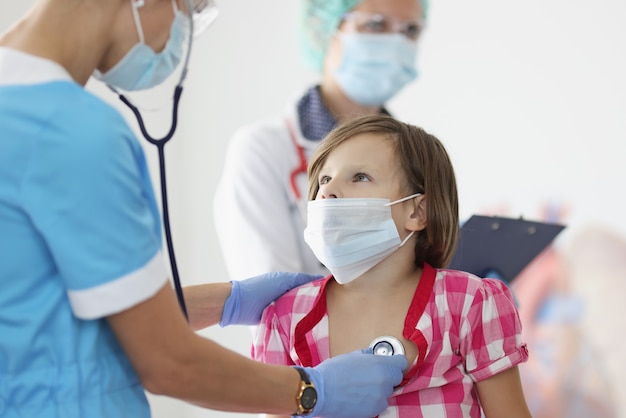 Foto doctor de máscara médica protectora escuchando corazón con estetoscopio a niña