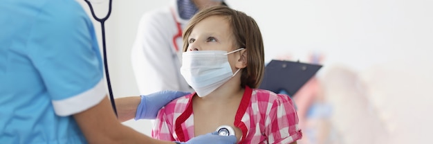 Doctor de máscara médica protectora escuchando corazón con estetoscopio a niña