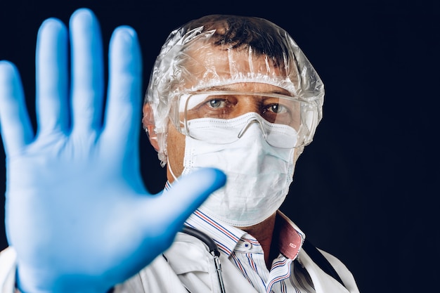 Doctor masculino mostrando señal de stop. Doctor vistiendo guantes y mascarilla médica. De cerca.