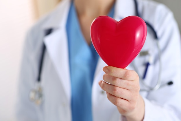 Doctor en medicina femenina manos sosteniendo un corazón de juguete rojo delante de su primer pecho. Ayuda médica, atención de cardiología, salud, profilaxis, prevención, seguros, cirugía y concepto de reanimación.