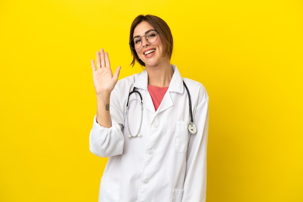 Doctor mujer aislada sobre fondo amarillo saludando con la mano con expresión feliz