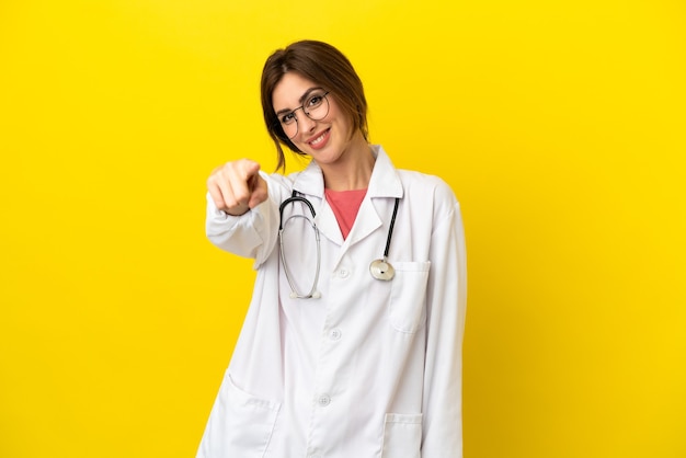 Doctor mujer aislada sobre fondo amarillo le señala con el dedo con una expresión de confianza