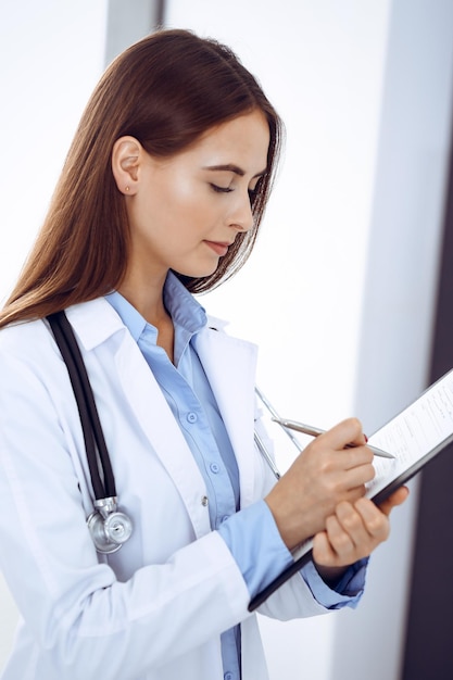 Doctor mujer llenando formulario médico mientras está de pie cerca de la ventana en la clínica. Médico en el trabajo. Concepto de medicina y atención de la salud.