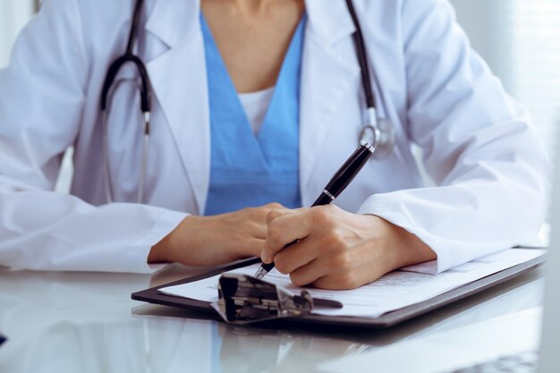 Doctor mujer llenando formulario médico mientras está sentado en la mesa, primer plano de las manos.