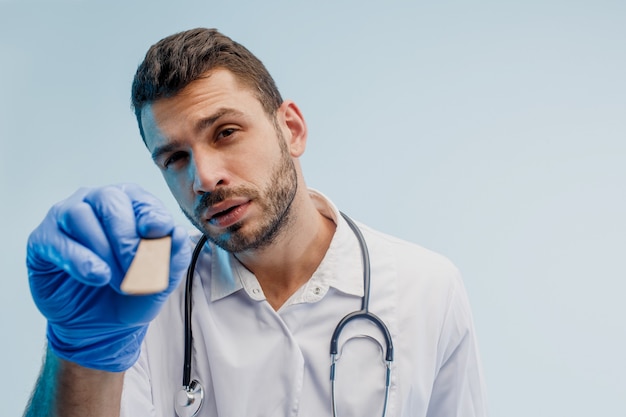 Doctor en otorrinolaringología masculino europeo enfocado con espátula médica. Hombre joven con estetoscopio con bata blanca y guantes de látex. Aislado sobre fondo gris con luz turquesa. Sesión de estudio