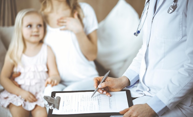 Doctor y paciente. Pediatra usando el clipboard mientras examina a una niña con su madre en casa. Feliz niño caucásico lindo en el examen médico. Concepto de medicina.