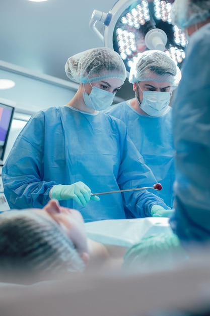 Doctor preocupado frunciendo el ceño mientras mira el estómago de un paciente en el quirófano. Trabajadora médica con algodón en fórceps