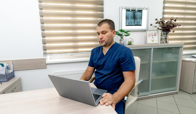 Doctor que trabaja con la computadora portátil en la oficina de la clínica. Médico en batas azules sentado en la mesa. Interior moderno en hospital.