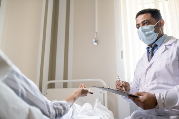 Doctor en ropa formal, bata blanca y máscara protectora tomando notas en un documento médico mientras escucha al paciente enfermo durante la conversación