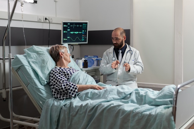 Doctor sentado al lado del paciente mayor enfermo tan enfermo tendido en la cama