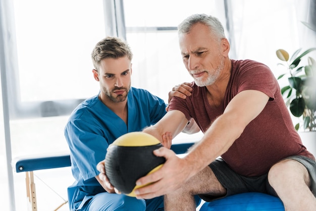 Doctor sentado cerca de un paciente maduro haciendo ejercicio en una pelota de fitness