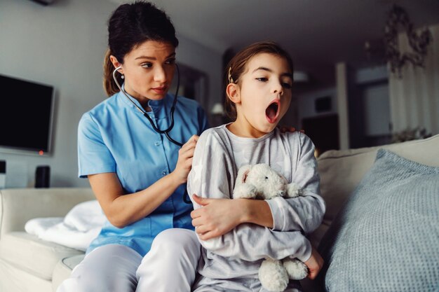 Doctor sentado en el sofá junto a la niña y examinando sus pulmones con estetoscopio.