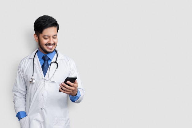 Doctor sonriendo y mirando el teléfono. Hombre pakistaní Doctor Tecnología Medicina en casa.