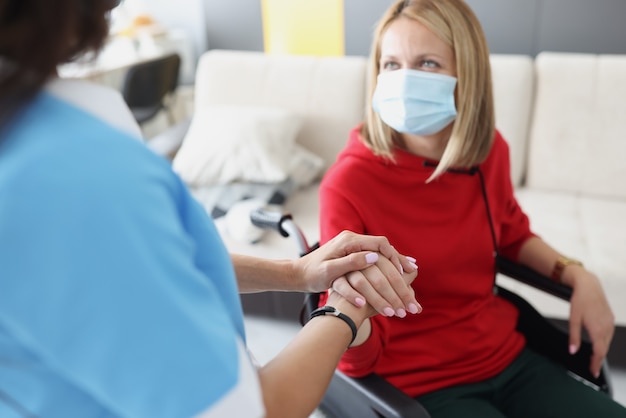 Doctor sosteniendo la mano de la mujer discapacitada en silla de ruedas en máscara médica protectora