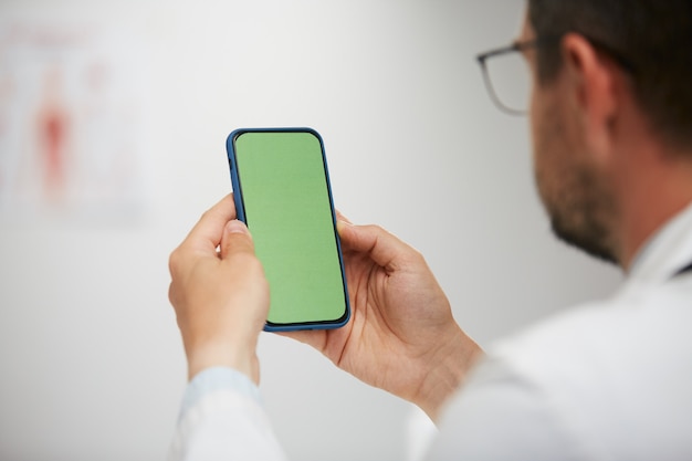 Doctor sosteniendo un teléfono inteligente de pantalla verde, médico senior con teléfono chroma key sentado en un gabinete médico con bata blanca y gases