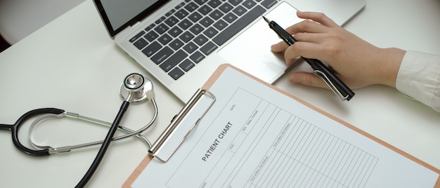 Doctor trabajando con computadora portátil, historia clínica y estetoscopio en la mesa de trabajo en la sala de examen