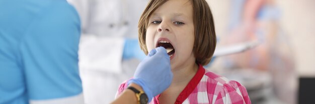 Doctor en traje azul mira la garganta del niño con un palo de madera.