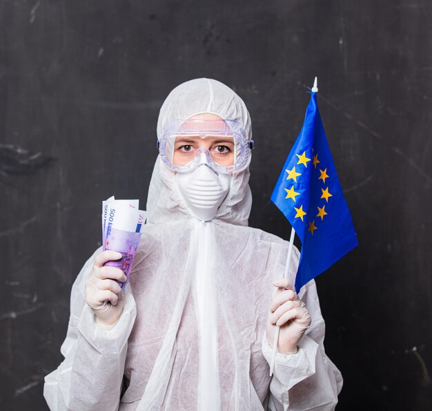 Foto doctor en traje de protección y gafas con máscara tiene dinero euro y bandera