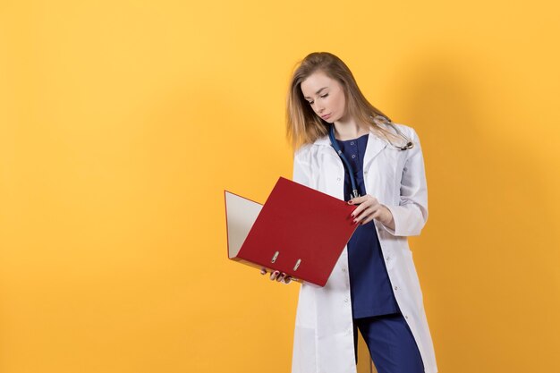 Doctor en traje quirúrgico y bata blanca sostiene una carpeta roja sobre un fondo amarillo