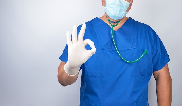 Doctor en uniforme azul y guantes estériles de látex muestra un símbolo de aprobación