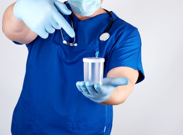 Doctor en uniforme azul y guantes de látex sostiene un recipiente de plástico vacío para tomar muestras de orina.