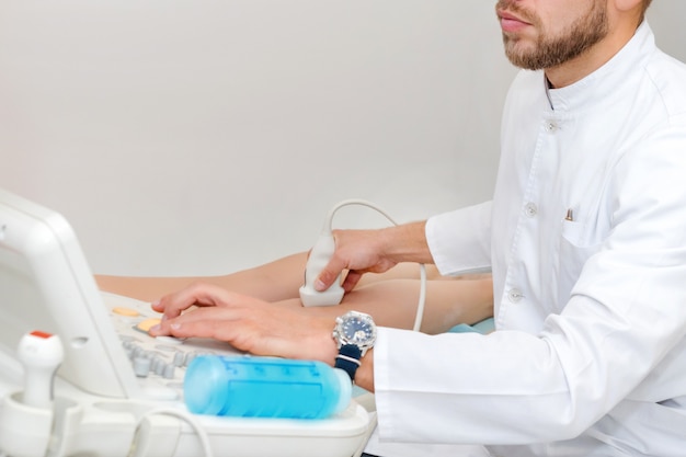 Doctor en uniforme blanco con equipo de diagnóstico realiza una ecografía utilizando un dispositivo en las piernas de una paciente. Hombre cirujano, flebólogo que trabaja en la clínica moderna. Herramientas médicas en el hospital.