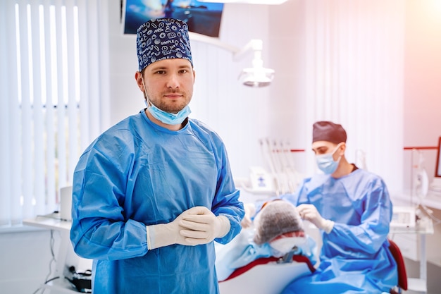 Doctor vestido con abrigo azul que lo invita a la oficina del dentista y mirando a la cámara con sillón estomatológico y paciente en el.