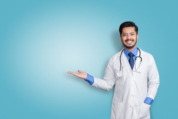 Doctor vistiendo uniforme sonriendo mientras presenta y apunta aislado en la pared azul con espacio de copia