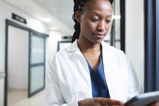 Doctora afroamericana con bata de laboratorio usando una tableta en el pasillo del hospital