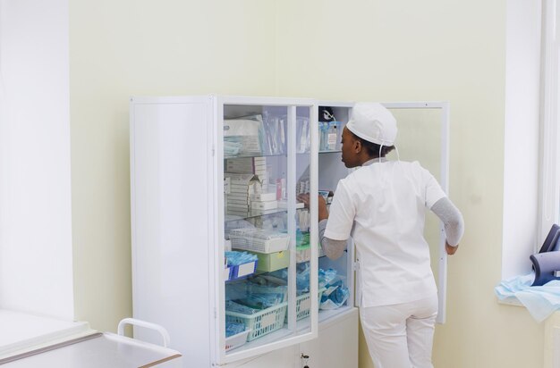 La doctora afroamericana elige medicamentos en un gabinete de vidrio en la sala de tratamiento