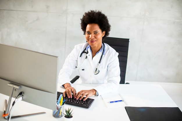 Doctora afroamericana vistiendo bata blanca con estetoscopio sentado detrás del escritorio en la oficina