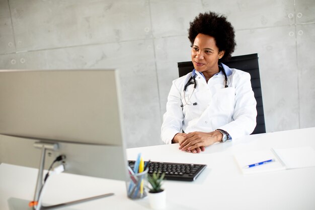 Doctora afroamericana vistiendo bata blanca con estetoscopio sentado detrás del escritorio en la oficina