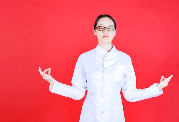 Doctora en anteojos de pie sobre fondo rojo y haciendo meditación.