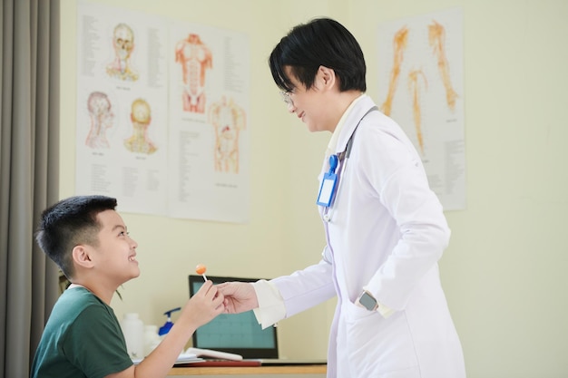 Una doctora asiática con bata blanca le da dulces a un niño mientras lo trata en el hospital