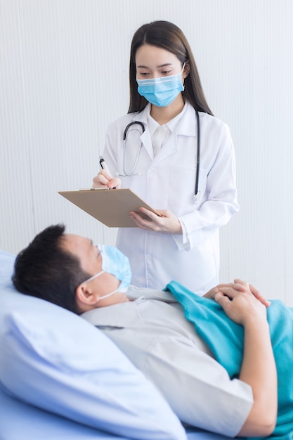 La doctora asiática está diagnosticando y registrando la salud de su paciente, que es un hombre asiático y se queda en la cama.