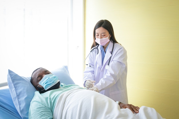 Una doctora asiática lleva guantes de inyección a un paciente afroamericano acostado en la cama. Concepto de servicio médico hospitalario