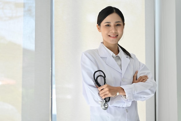 Doctora asiática profesional en bata blanca sosteniendo estetoscopio con los brazos cruzados