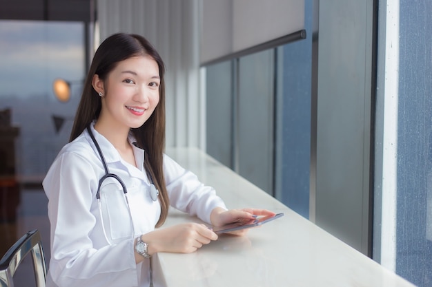 La doctora asiática profesional vestida con bata médica blanca se sienta en una silla en la biblioteca de un hospital para buscar información en una tableta para el tratamiento en el concepto de atención médica.