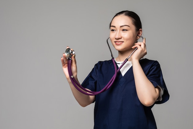 Doctora asiática sonriente alegre que examina con el estetoscopio, aislado sobre la pared blanca