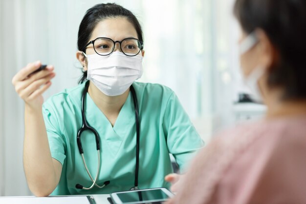 Foto doctora asiática en uniforme verde usar anteojos y máscara quirúrgica hablando, consultando y dando consejos a paciente anciana en sala médica en el hospital.