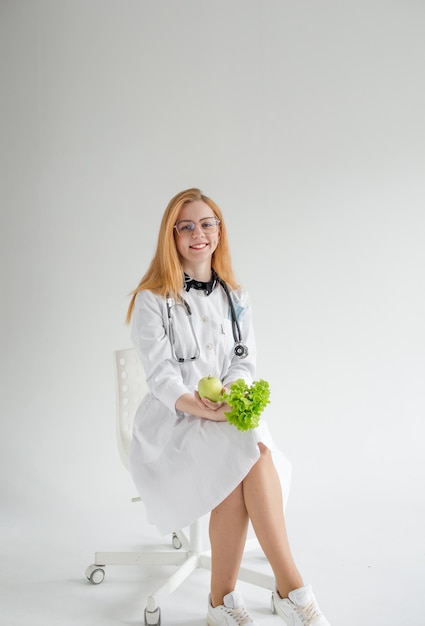 Una doctora atractiva con una bata blanca está sentada sonriendo y sosteniendo una manzana y una ensalada