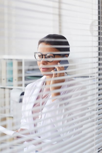 Doctora en bata blanca hablando por teléfono móvil mientras está de pie en la oficina detrás del obturador