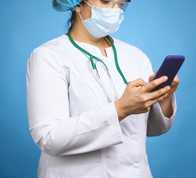 Doctora con bata blanca, máscara médica desechable y gafas sostiene un teléfono inteligente sobre un fondo azul.