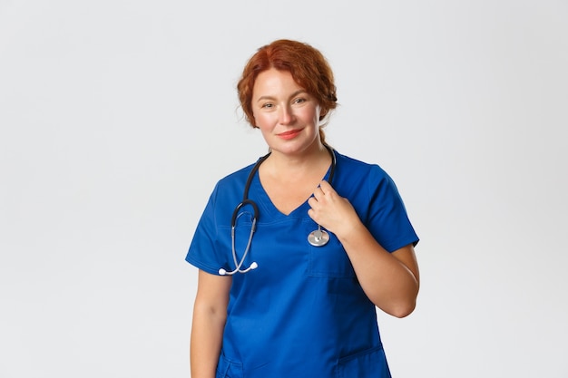 Doctora en batas azules con estetoscopio sonriendo a la cámara, tratando a los pacientes en el hospital de pie en gris