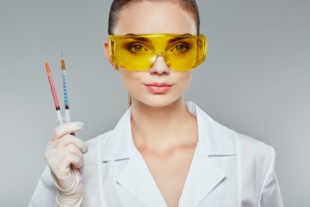 Doctora con cabello castaño y maquillaje desnudo vistiendo uniforme médico blanco, gafas, estetoscopios y guantes blancos en el fondo gris del estudio y sosteniendo jeringas, de cerca.