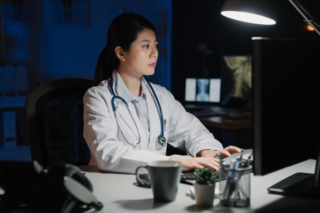 una doctora china asiática escribiendo en una computadora en una oficina oscura a medianoche. señora enfermera exceso de trabajo leyendo correo electrónico en línea en el lugar de trabajo de la clínica a altas horas de la noche. concepto de salud médica de trabajo de horas extra de personas