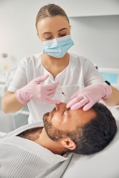 Doctora cosmetóloga en guantes estériles y mascarilla médica haciendo inyección en la frente masculina
